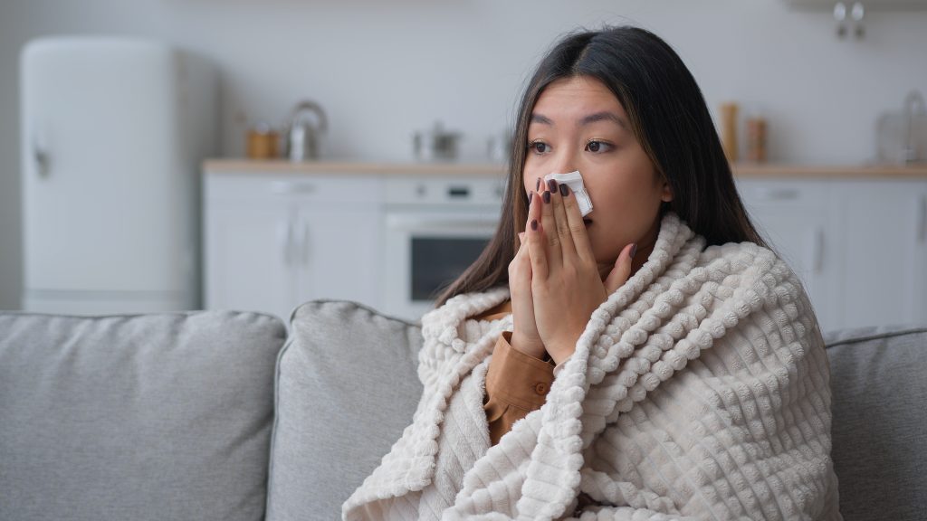 Unhealthy ill sick woman covered in warm blanket at home blowing nose from mold allergy